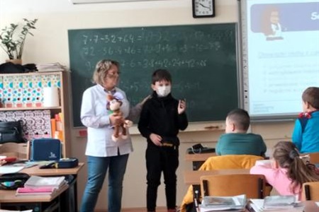 na środku klasy stoi pielęgniarka i uczeń, oboje ubrani w maseczkę, rozmawiają z resztą klasy