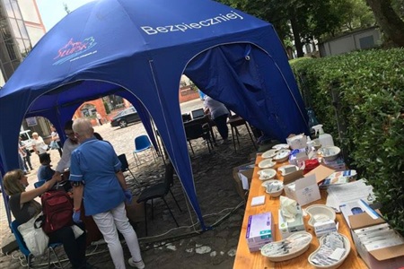 na pierwszy planie stół z asortymentem medycznym, w tle niebieski namiot pod którym pielęgniarka wykonuje szczepienie 