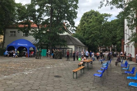 brukowany dziedziniec, w oddali pod drzewami niebieski namiot do którego czekają pacjenci ustawieni w kolejkę, z przodu niebieskie krzesełka i drewniane stoły