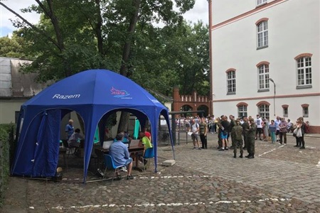 grupa żołnierzy w mundurach czaka przed niebieskim namiotem, w tle budynek i zgromadzeni przed nim ludzie