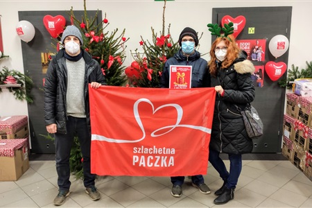 kobieta i dwóch mężczyzn na tle choinki prezentują czarowny banek z napisem szlachetna paczka