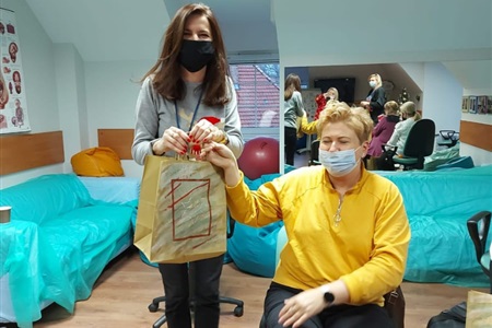 kobieta siedzi na krześle, koło niej stoi kobieta z torbą prezentową w ręku
