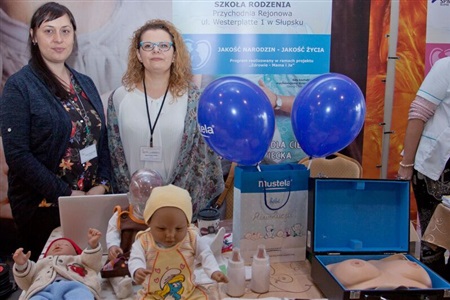 dwie kobiety stoją, przed nimi na stoliku leżą manekiny dzieci, 2 butelki na mleko, laptop oraz model piersi 