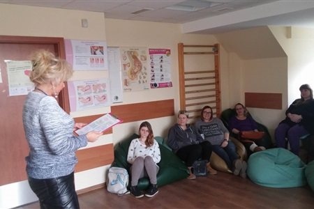 grupa siedzących na pufach kobiet słucha stojącego przed nimi kobiety