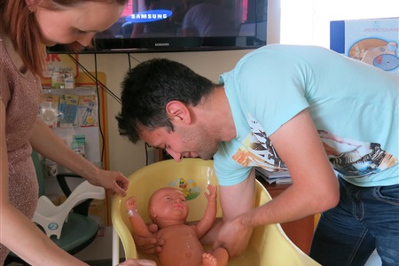 mężczyzna nachylony nad wanienką, symuluje kąpiel dziecka trzymając manekina, towarzyszy mu stojąca obok kobieta