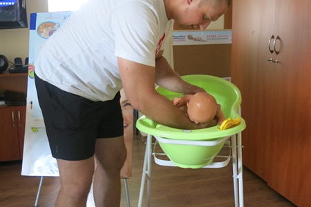 mężczyzna nachylony nad wanienką, symuluje kąpiel dziecka trzymając manekina